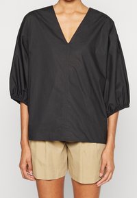 Blusa nera leggera con scollatura a V, maniche ampie e gonfie, e una vestibilità morbida, abbinata a pantaloni shorts beige. Texture liscia, design minimalista.