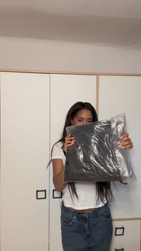 Articolo confezionato in nero avvolto in plastica parzialmente trasparente, con superficie liscia. Sullo sfondo si vedono armadi bianchi con dettagli neri.