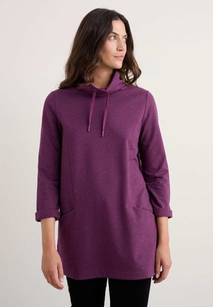 Tunique à capuche violette avec des manches longues, présentant un col croisé et deux poches avant. Fabriquée dans un tissu doux et texturé.