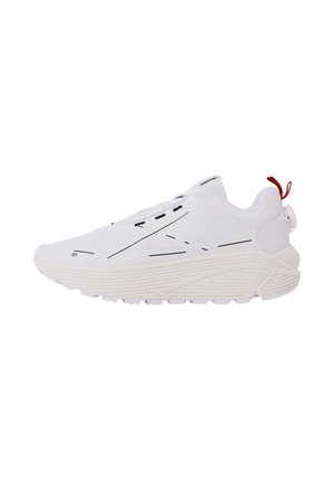 Baskets de sport blanches avec une semelle épaisse texturée, des accents de lignes noires minimalistes et une languette rouge au talon.