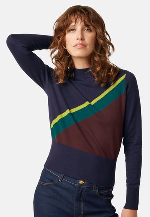 Mujer con cabello rizado y castaño lleva un suéter azul marino con rayas diagonales verdes, amarillas y marrones y vaqueros azul oscuro, posando con un brazo levantado.