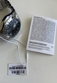 Sølv rustfrit stål ur med en sort struktureret urskive, chronograf funktioner, og et hvidt garantimærke fastgjort med en snor.