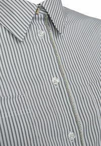 Camisa de botones a rayas en gris y blanco. Presenta un collar, dos bolsillos frontales y una cremallera decorativa con acentos de perlas a lo largo de la tapeta.