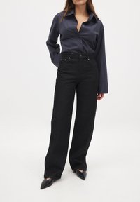 Mustad kõrge vöökohaga mustad teksad, millel on laia säärefit, valmistatud denimist. Kombineeritud tumedast hallist nööbiga särgist ja terava ninaga mustadest kingadest.