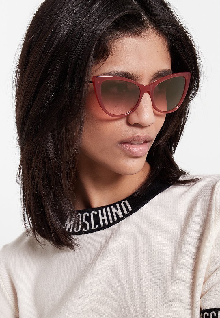 Occhiali da sole cat-eye rossi con lenti sfumate, caratterizzati da una montatura spessa e una texture liscia. Il modello indossa una camicia con un colletto con logo nero.