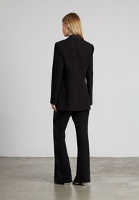 Blazer nero su misura con un design aderente, abbinato a pantaloni neri a sbuffo con dettagli in pizzo texture lungo il lato.