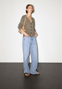 Brązowy, zapinany na guziki kardigan z teksturowanym wzorem, połączony z jasnoniebieskimi spodniami typu wide-leg i czarnymi klapkami na neutralnym tle.