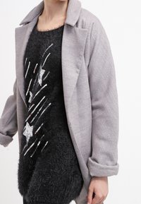 Abrigo estructurado gris con cuello de muesca, llevado sobre un suéter negro y esponjoso que presenta un patrón de estrellas plateadas y líneas diagonales.