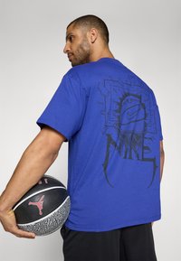 Violet Nike marškinėliai su juodu grafikiniu dizainu ant nugaros, vaizduojančiu krepšinio kamuolį ir kampuotas akcentus, su paprastu priekiu.