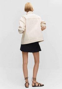 Veste en jean beige oversize avec des manches retroussées, présentant un design ample. Associée à une mini-jupe noire et des sandales noires à lanières.