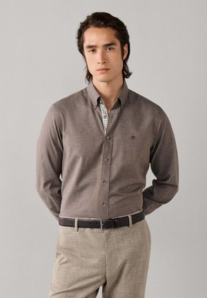 Hombre de pie con las manos detrás de la espalda, vistiendo una camisa marrón abotonada, camiseta de cuadros, cinturón marrón y pantalones beiges a cuadros sobre un fondo liso.
