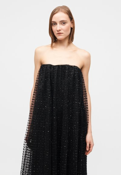 Woman with straight light brown hair wears strapless black dress with textured sheer overlay, standing against plain light background.