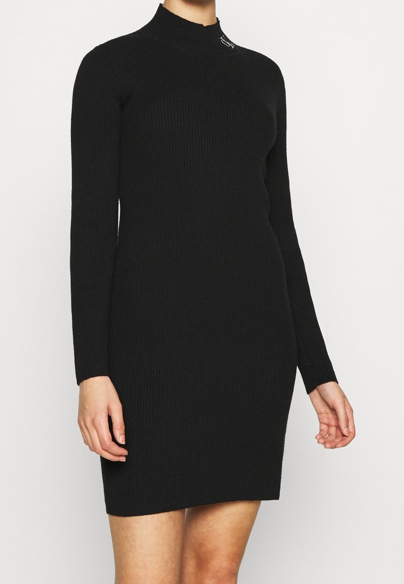 Femme portant une robe noire en maille côtelée, ajustée, à manches longues, col montant et ourlet à hauteur du genou, debout devant un fond uni.