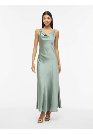Kvinne iført en lys grønn sateng ermeløs maxikjole med drapert utringning og beige flettede heeled sandaler, stående mot hvit bakgrunn.