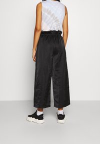 Person med svarta vida kapade byxor, vita strumpor, svarta sneakers och en ärmlös tie-dye-topp, vänd mot en enkel ljus bakgrund.