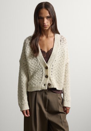 Woman wearing a white knitted buttoned cardigan over a brown lace top and loose brown trousers, standing against a plain background.