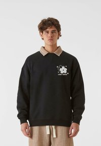 Melna sporta džemperis ar beigtu apkakli, kurā ir balts zieda grafiskais attēls un uzraksts "ZUDUSĪ BŪTNE". Uzvilkts virs rakstainām beigtu biksēm.