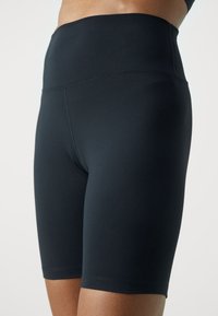 Obcisłe, czarne rowerowe szorty wysokiej talii noszone przez modelkę, sięgające do połowy uda, z widocznymi detalami szwów.