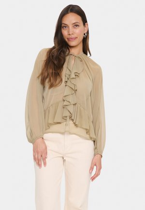 Light khaki blouse with ruffled design, tie neckline, and long sleeves. Fabric appears lightweight and semi-sheer. Worn with light-colored pants.