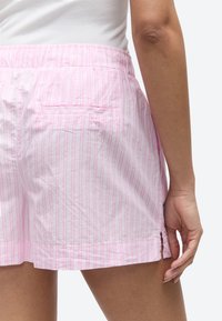 Moteris dėvi šviesiai rožines ir baltas dryžuotas šortus su galine kišene ir šoniniu plyšiu, derinančią su balta palaidine.