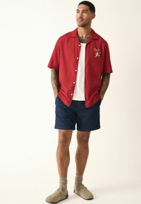Homme se tenant debout avec les mains dans les poches, portant une chemise à boutons à manches courtes rouge, un débardeur blanc, un short bleu marine, des chaussettes beiges et des chaussures slip-on beiges.