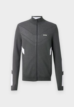 Graue Zip-Jacke mit hohem Kragen, die über ein strukturiertes Design am oberen Oberkörper und weiße Akzente an den Ärmeln verfügt. Logo auf der Brust.