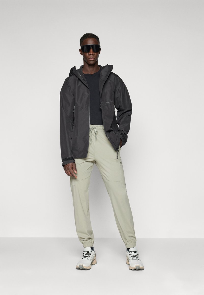 Giacca impermeabile nera con cappuccio, abbinata a pantaloni elastici verde chiaro con polsini. Il modello indossa occhiali da sole neri e scarpe bianche.