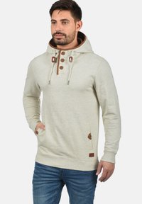 Lichtgrijs hoodie met een gestructureerde afwerking, bruine koorden, knoopaccenten en zijzakken. Draagt men met blauwe spijkerbroeken.
