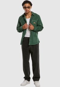 Grønn cordfløyelskjorte med brystlomme og logo, kombinert med svarte jeans og hvite sneakers. Modellen har solbriller og en singlet på.