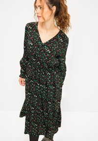 Vestido negro con un patrón abstracto en verde, rojo y blanco, que presenta un escote en V, mangas largas y un diseño fluido hasta los tobillos.