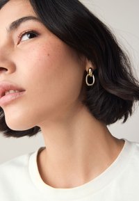 Gold-tone hoop earrings with an interlocking design, featuring a polished finish. Hair is dark with a wavy texture, and the shirt is off-white.