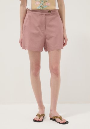 Persoon draagt stoffig roze shorts met hoge taille, een knoop aan de voorkant en zakken, gecombineerd met bruine platte sandalen met studs en een top met bloemenprint.