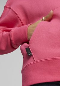 Sudadera rosa con tejido texturizado, que cuenta con un bolsillo frontal y una pequeña etiqueta negra de Puma cosida en la costura lateral.