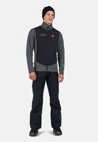 Svart och grå isolerad väst med hög krage, dragkedjestängning och två sidofickor. Bärs med svarta byxor och en svart mössa.