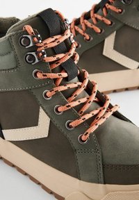 Chaussures montantes vertes en tissu texturé avec des panneaux lisses. Équipées de lacets à motifs orange et noir, d'œillets en métal argenté et de semelles beiges.