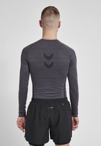 Langærmet atletisk t-shirt i mørkegrå med struktureret stof og reflekterende chevronmønster på ryggen; parret med sorte atletiske shorts.