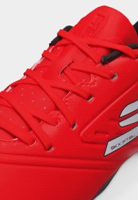 Κόκκινο αθλητικό παπούτσι με κορδόνια, μαξιλαρωμένη γλώσσα, οπές για αερισμό και λευκή ετικέτα "SKX:FTBL" στο πλάι.