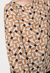 Blusa con motivo geometrico nei toni del marrone, crema e nero. Tessuto morbido con maniche a pieghe, scollatura tonda e texture visiva.
