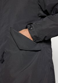 Black waterproof jacket featuring a smooth texture, adjustable drawstring hem, and a side pocket with a button closure.