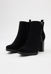 Botas de tobillo de ante negro con tacón grueso, cremallera lateral y tirador en la parte superior; textura suave con un acabado mate y suela de goma.
