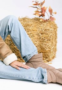 Stivaletti alla caviglia in suede beige con dettagli traforati, indossati con jeans di denim azzurro chiaro. L'outfit include una giacca beige foderata in pile.