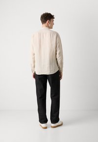 Camicia in lino beige chiaro con orlo arrotondato e polsini con bottoni, abbinata a pantaloni in denim scuri e sneakers bianche, vista da dietro.