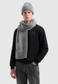Grijze geribbelde gebreide beanie en bijpassende sjaal, met geborduurd detail. Draagt over een zwarte sweatshirt en grijze jeans. Eenvoudig en gestructureerd ontwerp.