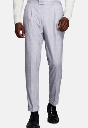 Man wearing light gray pinstripe trousers with cuffed hems, black dress shoes, and a white long-sleeve knit top, standing on a white background.
