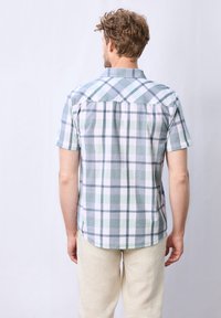 Košile s krátkým rukávem v kostkovaném vzoru v světle zelené, bílé a modré barvě. Má límec na knoflíky a detail na zadním díle. Nosit s krémovými kalhotami.