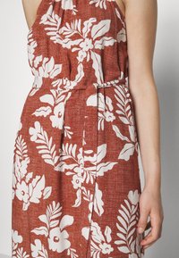 Robe sans manches couleur rouille avec motifs floraux et feuilles blancs, ceinture à la taille, gros plan du bras et de la main droite.