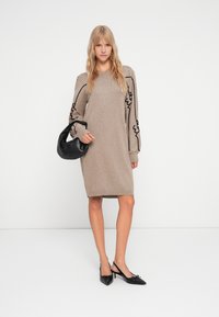 Robe en tricot beige à manches longues avec des coutures décoratives noires. Le mannequin tient un sac à main noir et porte des escarpins noirs à bout pointu.