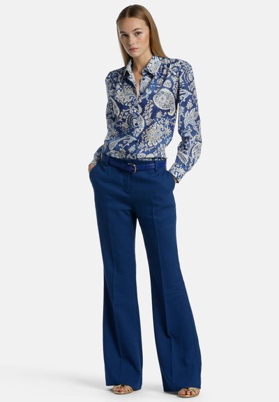 Langsleeve blauwe blouse met een witte paisleyprint, gecombineerd met hooggetailleerde blauwe broek. De broek heeft een wijde pijp en een riem.