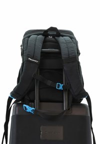 Mochila negra con correas acolchadas, clips azules, apoyada sobre una maleta negra. Tiene una superficie texturizada y una asa en la parte superior.