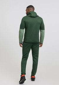 Ensemble de sport pour homme en vert foncé et vert clair, comprenant une veste à capuche et un pantalon fuselé. Le matériau est extensible et lisse.